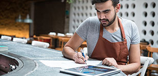 A young guy in a business on a tablet and writing down information on paper.